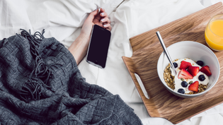  Hand coming out from under blanket with cell phone held loosely next to wooden tray with cereal and orange juice | Style Standard